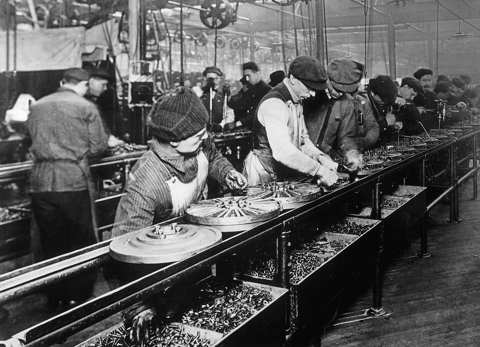 La catena di montaggio della Ford a Highland Park nel 1913: operai in fila lungo un banco su cui scorrono i volani dei motori, ognuno ripete lo stesso gesto